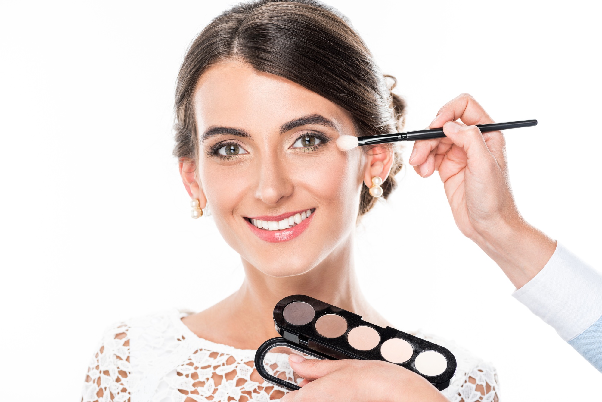 smiling woman looking at camera while makeup artist applying blush isolated on white